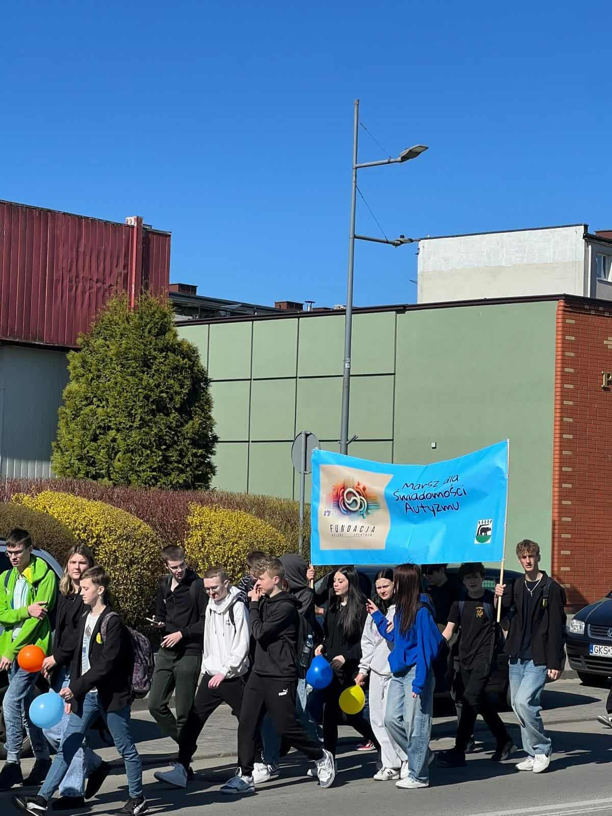 Uczestnicy marszu, głównie nastolatkowie, idący chodnikiem na tle miejskich budynków. Na czele niesiony jest duży błękitny baner z logiem fundacji i hasłem "Marsz dla Świadomości Autyzmu". Niektórzy uczestnicy trzymają w rękach niebieskie, żółte i pomarańczowe balony.