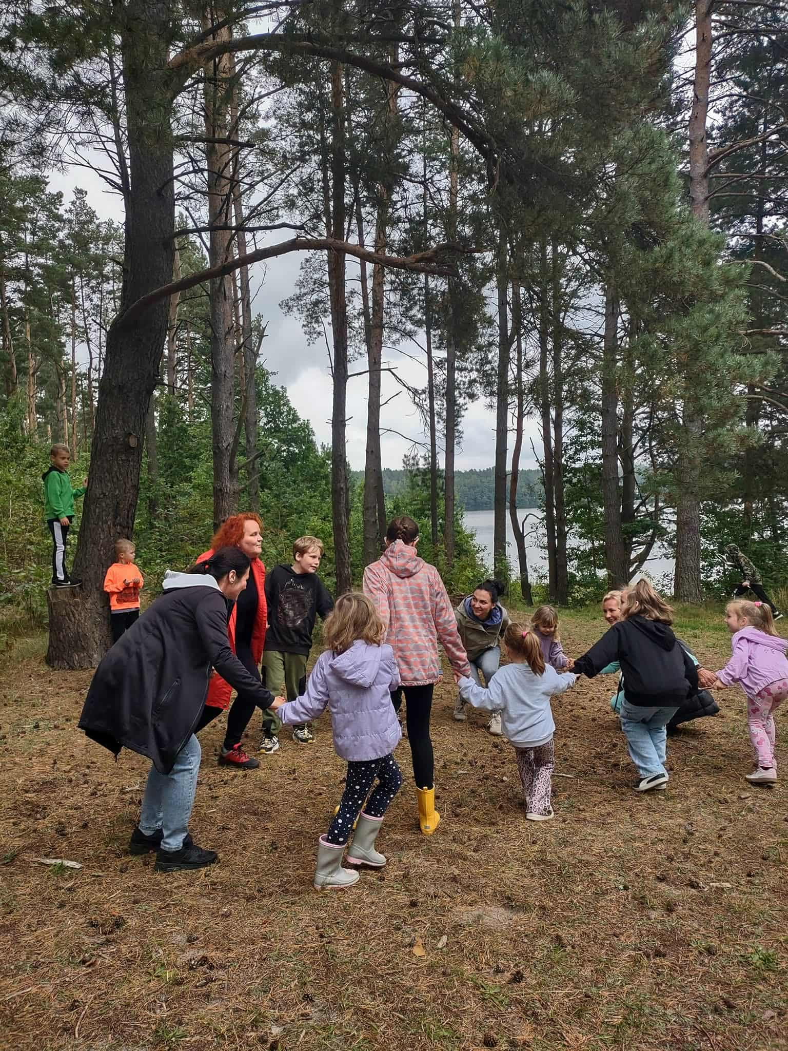 Grupa dzieci i dorosłych bawiąca się w kole, trzymając się za ręce w lesie sosnowym nad jeziorem.