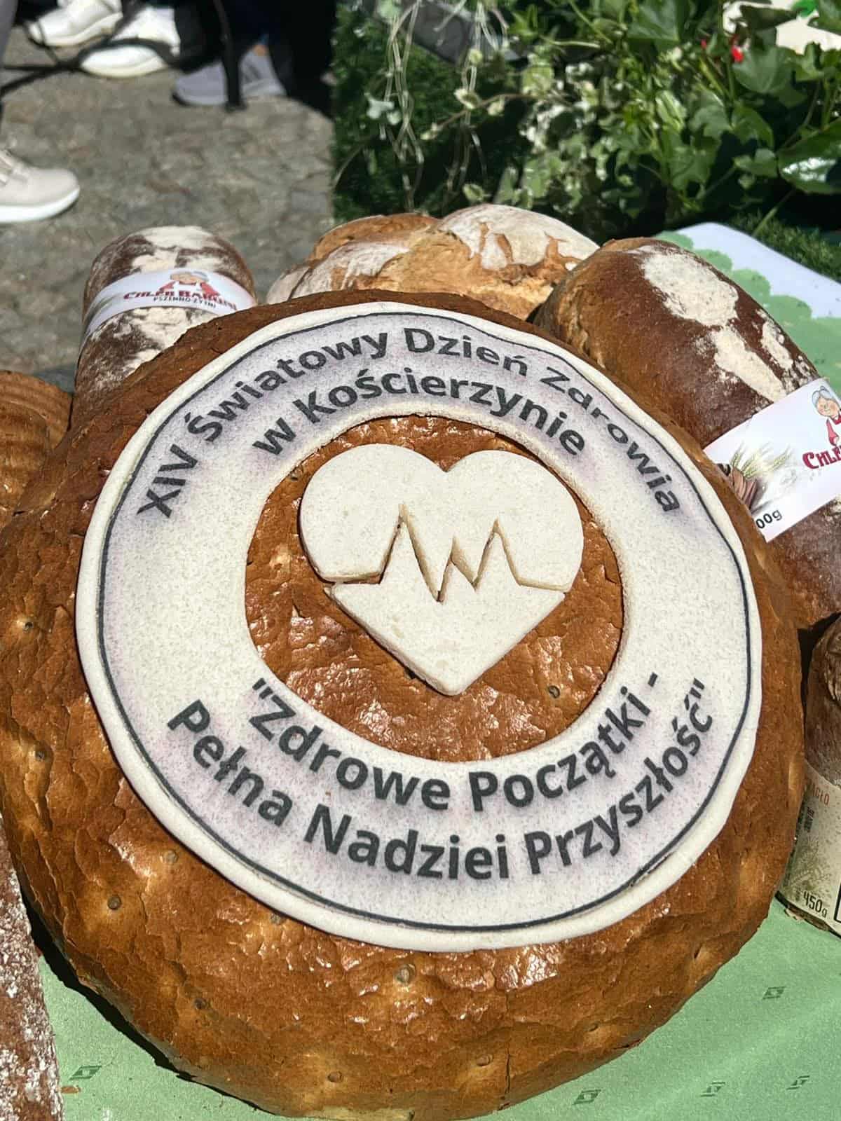 Okrągły bochenek chleba udekorowany napisem „XIV Światowy Dzień Zdrowia w Kościerzynie” oraz hasłem „Zdrowe Początki – Pełna Nadziei Przyszłość”. Na środku znajduje się symbol serca z linią EKG.