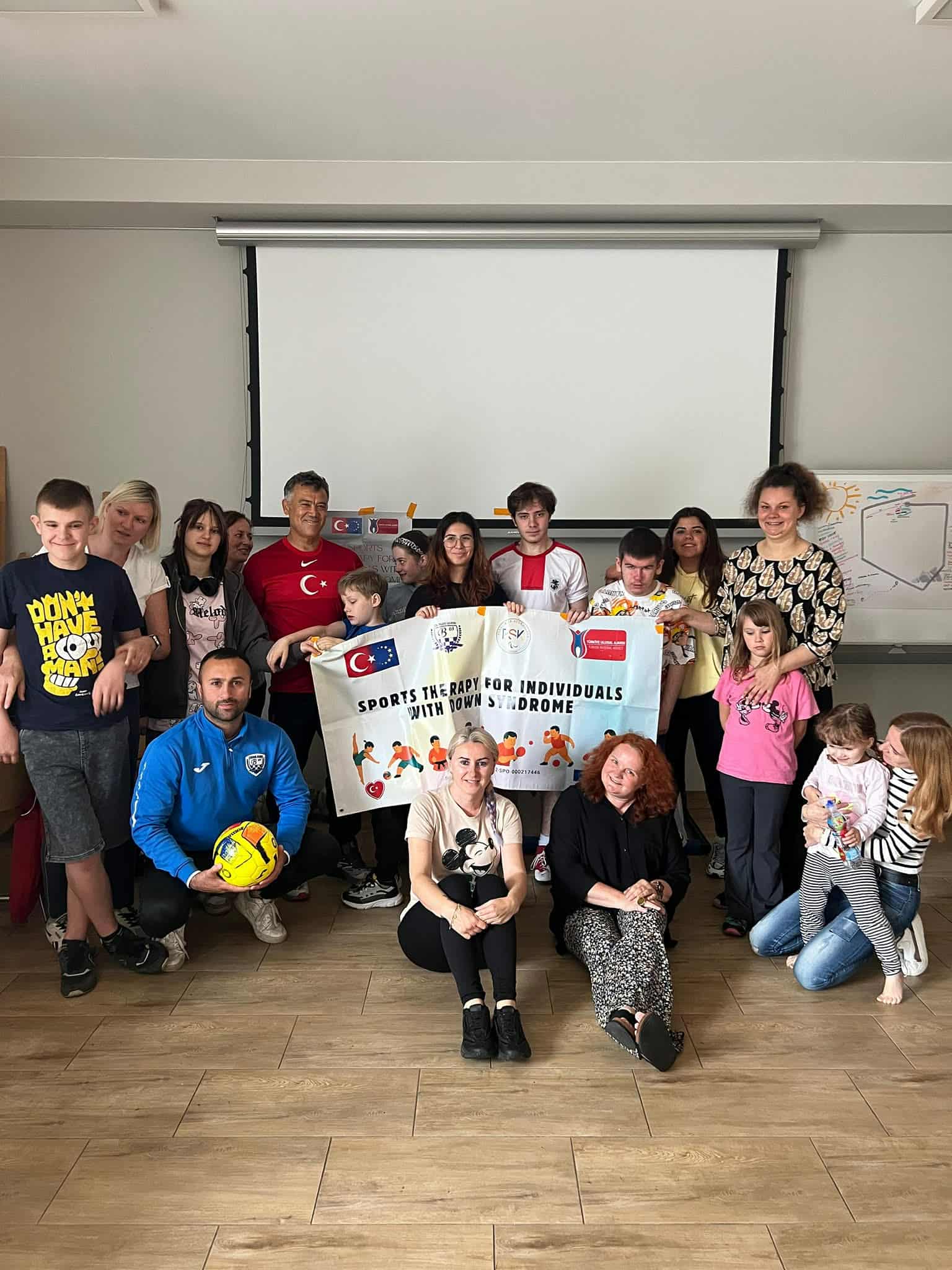 Grupa osób pozuje do wspólnego zdjęcia, trzymając baner z napisem „SPORTS THERAPY FOR INDIVIDUALS WITH DOWN SYNDROME”.