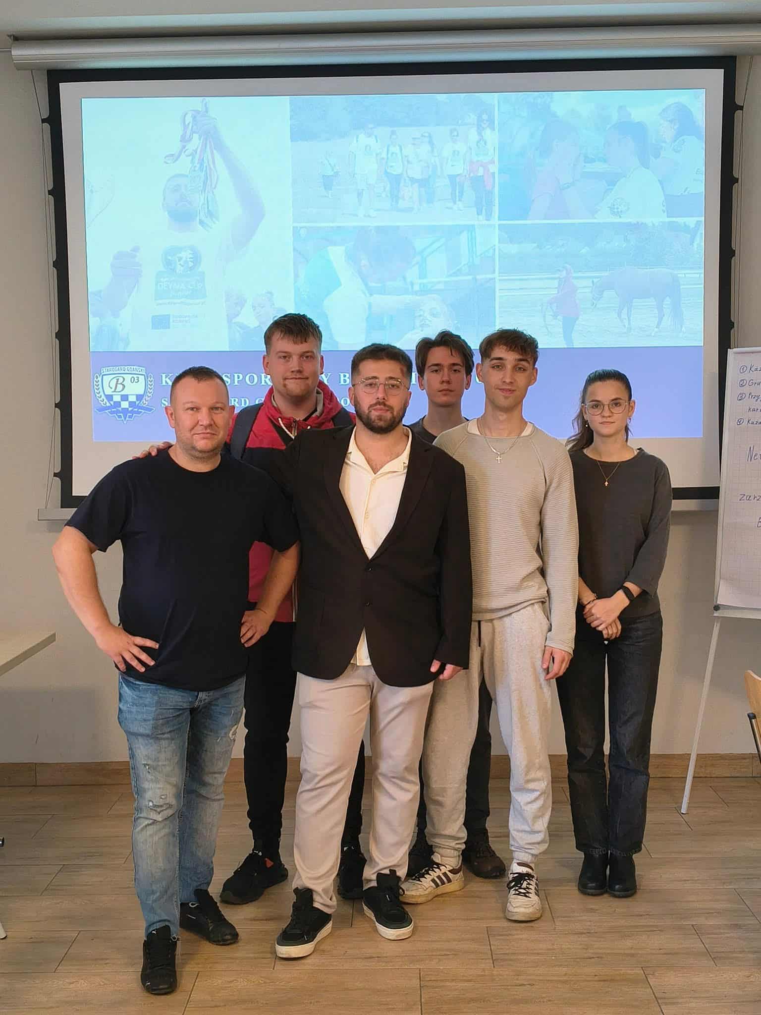 Grupa sześciu osób pozuje do pamiątkowego zdjęcia na tle ekranu projekcyjnego z logiem klubu sportowego KS Beniaminek 03 Starogard Gdański.